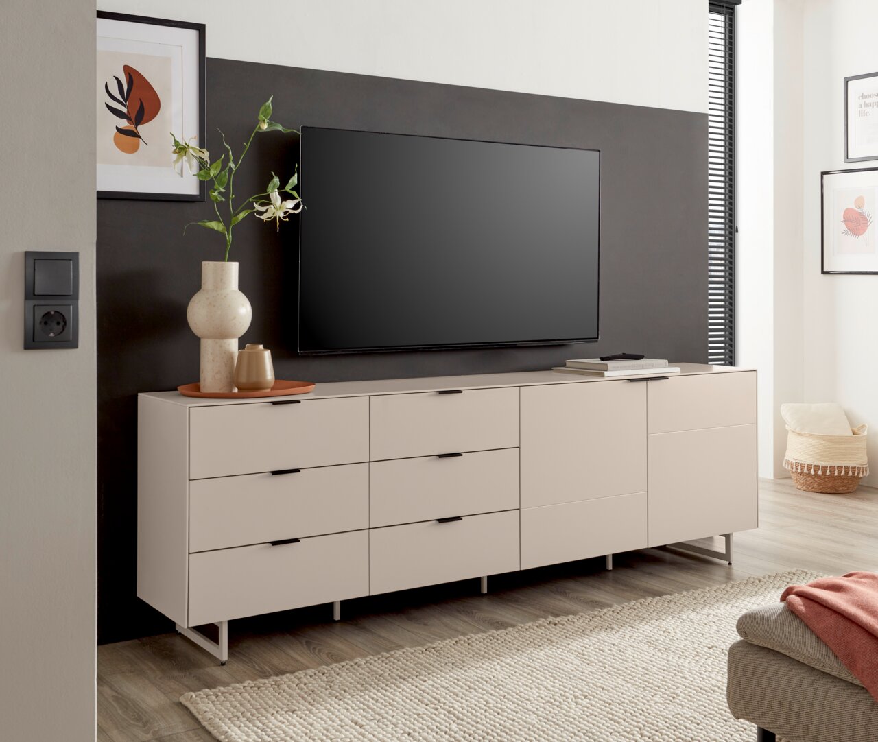 Modernes, hellbeiges TV-Lowboard mit Schubladen und Türen auf schlanken Metallfüßen, mit Fernseher und Deko im Wohnzimmer, frontale Perspektive.