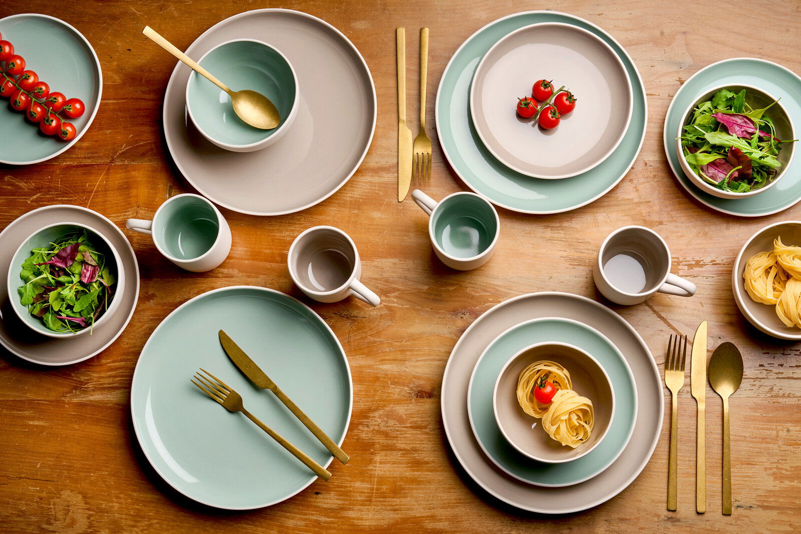 Kombiservice 16-tlg. in Vogelperspektive, bestehend aus Tellern, Schalen und Tassen in sanften Pastelltönen, arrangiert auf einem Holztisch mit goldfarbenem Besteck.