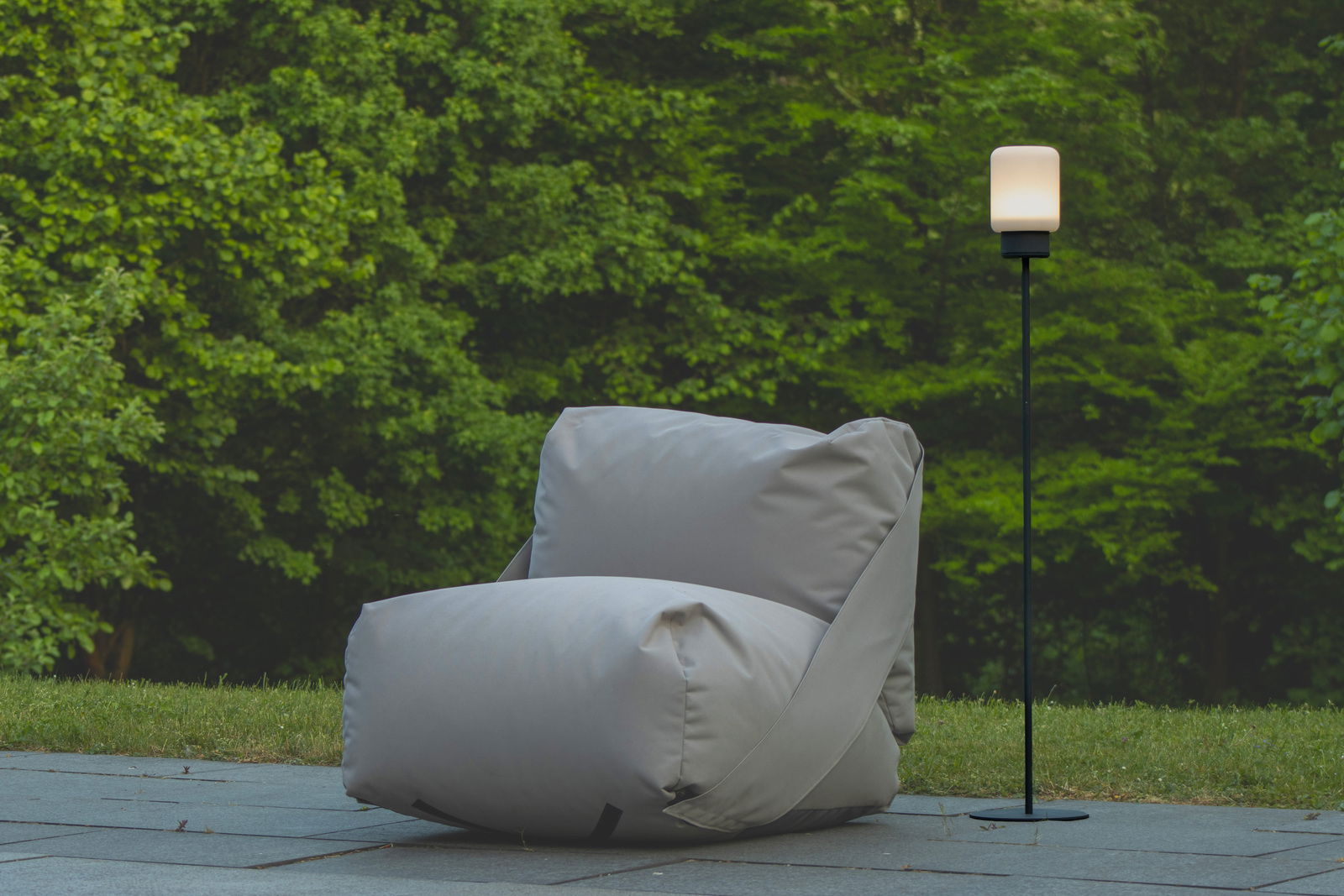 Ein modernes Akku-Windlicht auf einem schlanken schwarzen Gestell, im Freien neben einem großen grauen Sitzsack, mit grüner Natur im Hintergrund. Perspektive: seitlich.