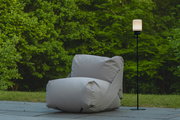 Ein modernes Akku-Windlicht auf einem schlanken schwarzen Gestell, im Freien neben einem großen grauen Sitzsack, mit grüner Natur im Hintergrund. Perspektive: seitlich.