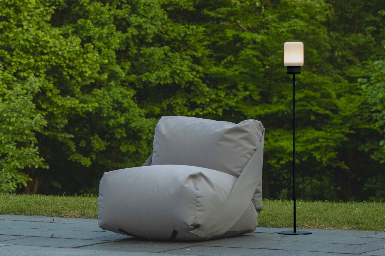 Ein modernes Akku-Windlicht auf einem schlanken schwarzen Gestell, im Freien neben einem großen grauen Sitzsack, mit grüner Natur im Hintergrund. Perspektive: seitlich.
