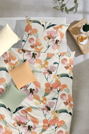 Bettwäsche-Set mit großflächigem Aquarell-Blumenmuster in Orange, Rosa und Grün auf weißem Stoff, Draufsicht von oben auf das gemachte Bett