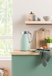 Isolierkanne Skyline in sanftem Grün auf einer Küchenarbeitsplatte, umgeben von Holzbrett, Geschirr und Pflanzen, seitliche Perspektive.