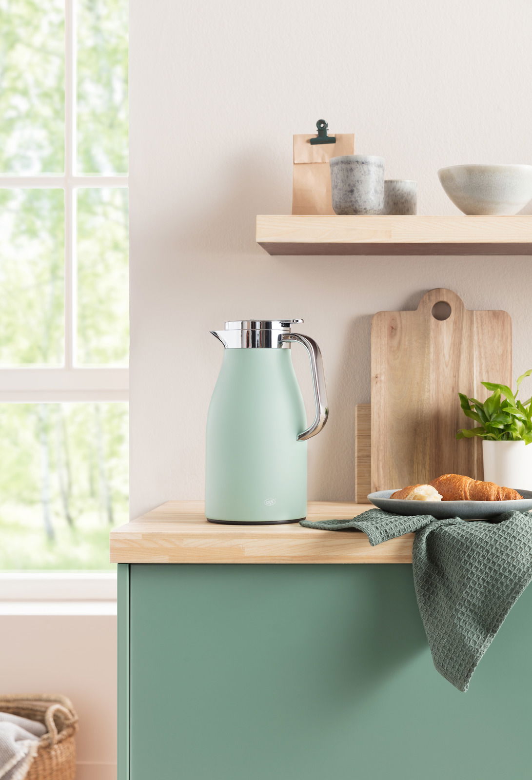 Isolierkanne Skyline in sanftem Grün auf einer Küchenarbeitsplatte, umgeben von Holzbrett, Geschirr und Pflanzen, seitliche Perspektive.