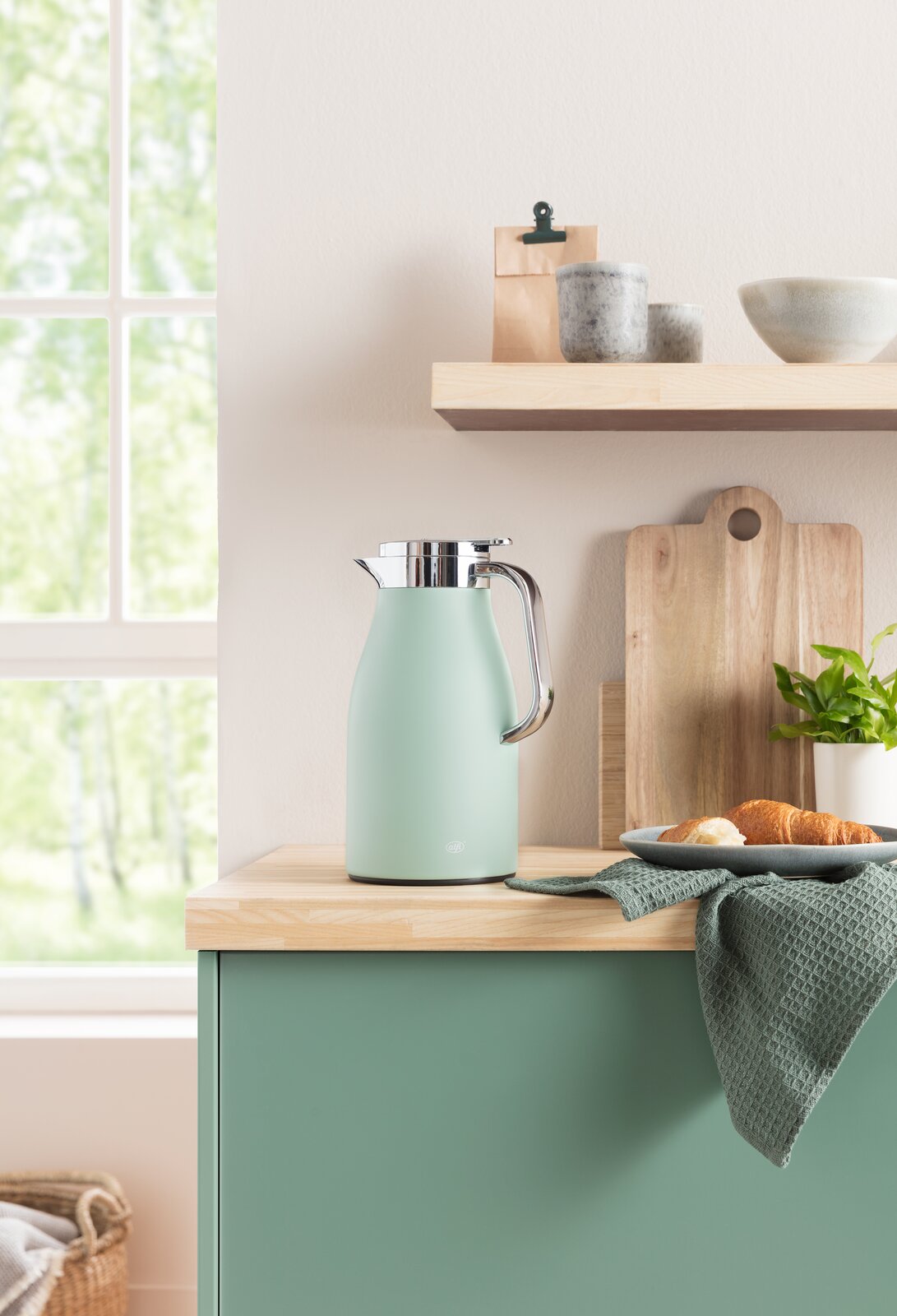 Isolierkanne Skyline in sanftem Grün auf einer Küchenarbeitsplatte, umgeben von Holzbrett, Geschirr und Pflanzen, seitliche Perspektive.