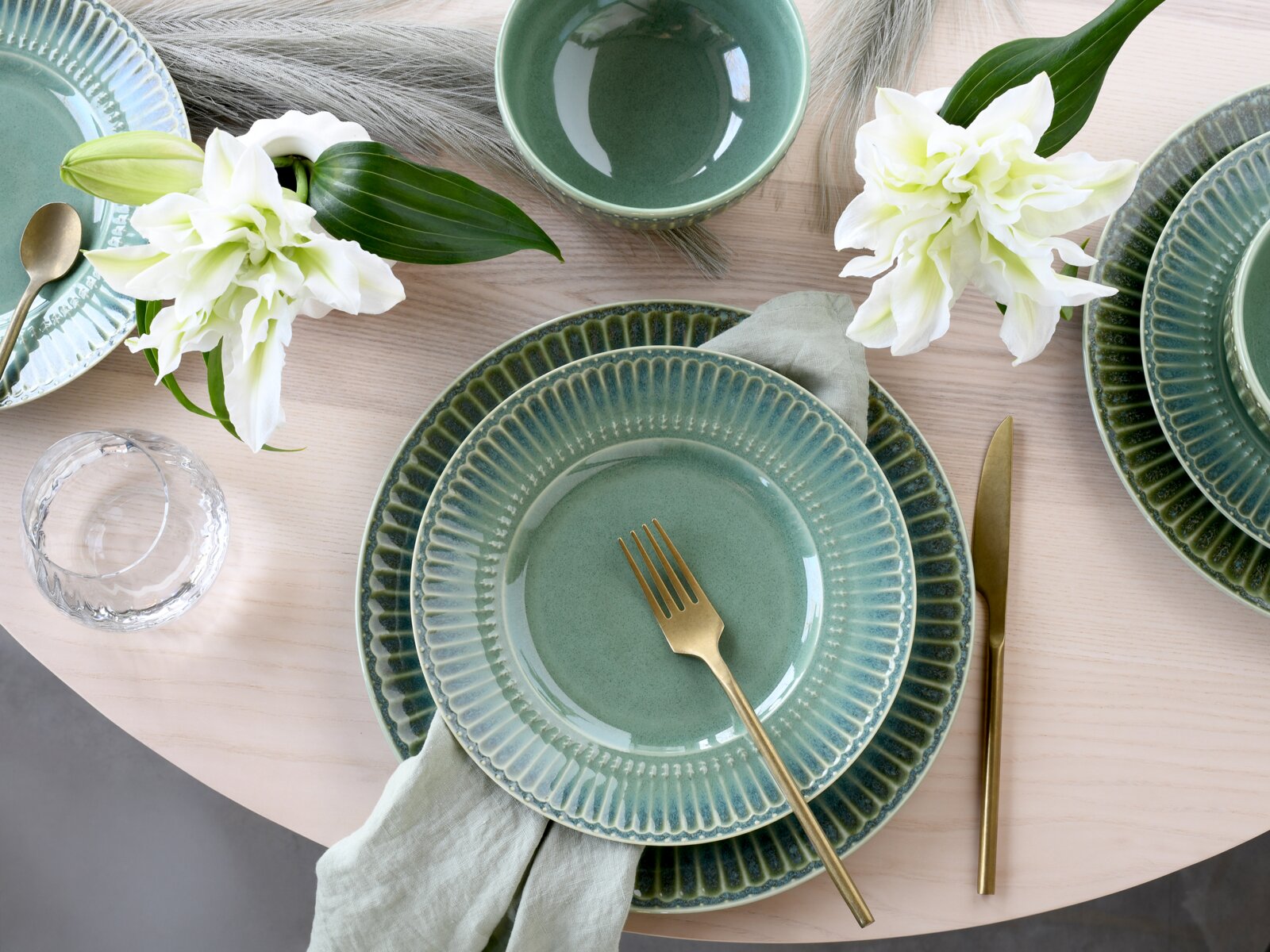 Draufsicht auf ein stilvoll gedecktes Tisch-Setting mit grün glasiertem Keramikgeschirr (Teller und Schale), goldfarbenem Besteck, Leinenserviette, Wasserglas und weißen Lilien auf hellem Holztisch.
