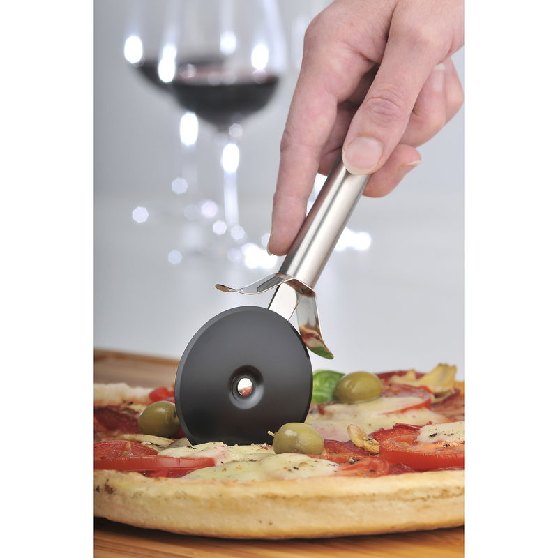 Nahaufnahme eines Pizzaschneiders Profi Plus, der eine Pizza mit Oliven und Tomaten schneidet. Im Hintergrund ist ein Glas Rotwein zu sehen.