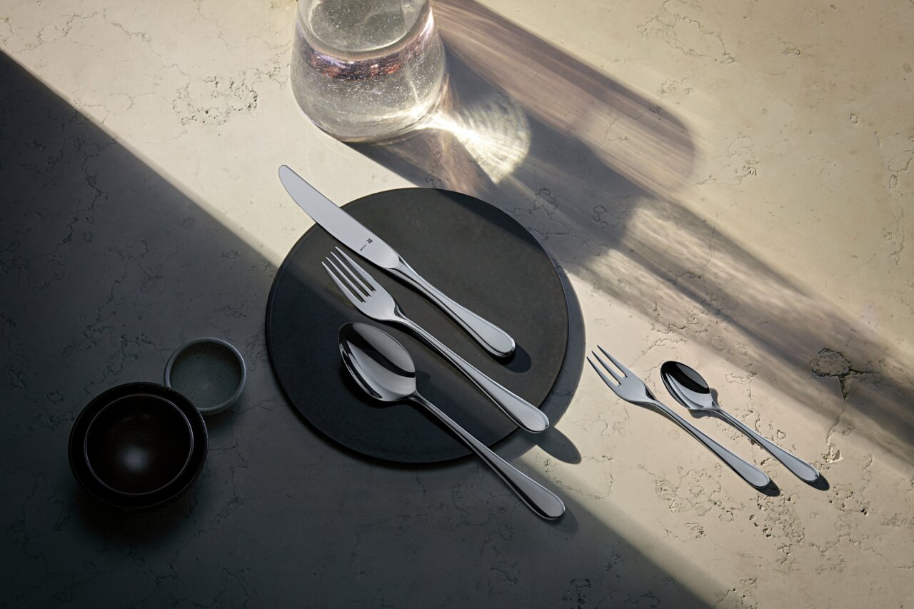Elegantes Besteckset auf einem dunklen Teller, bestehend aus Messer, Gabel und Löffel, in einer stilvollen Tischdekoration von oben betrachtet.