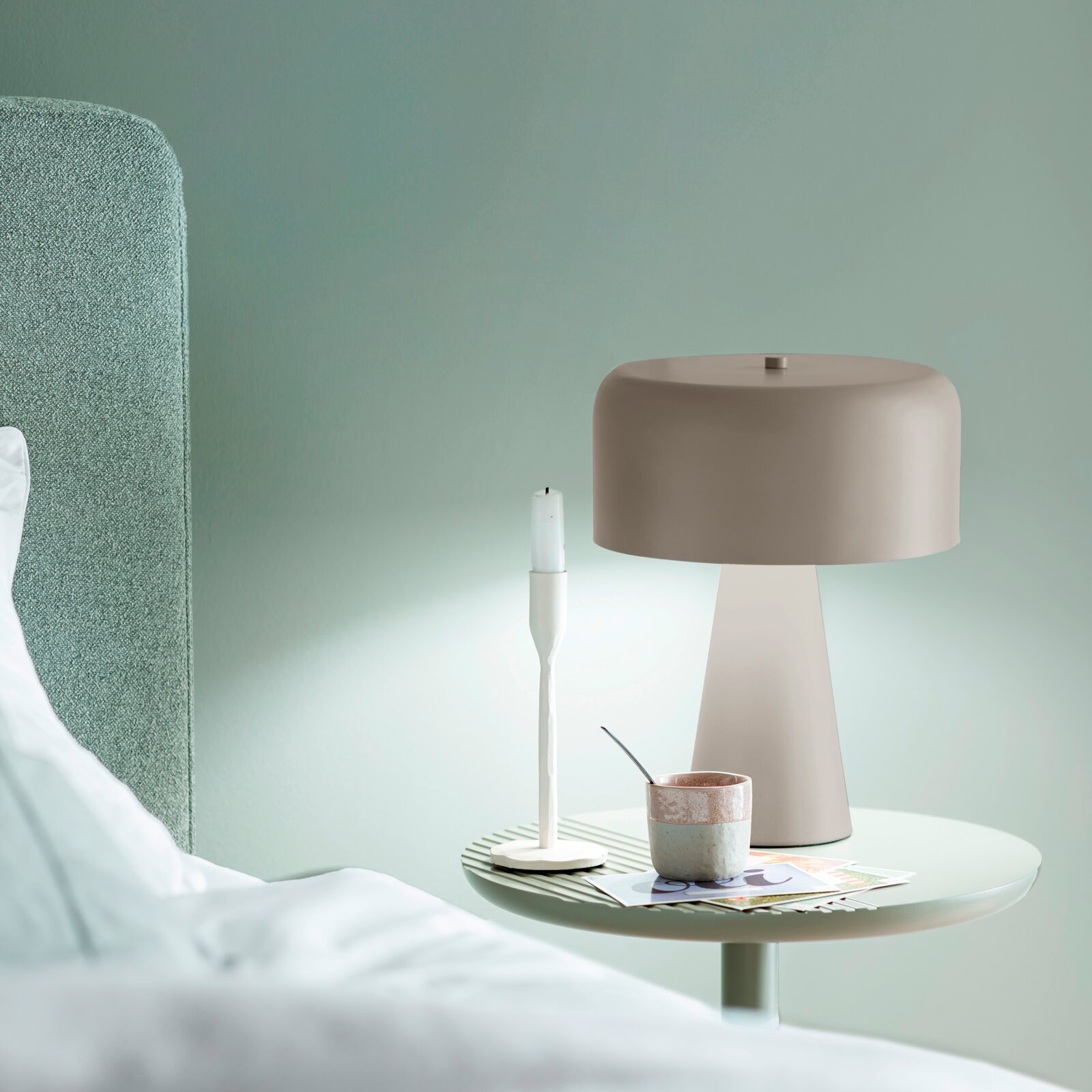 Beige Tischlampe mit breitem, rundem Schirm auf rundem Nachttisch neben Kerzenhalter und Tasse, Innenraumaufnahme in seitlicher Perspektive
