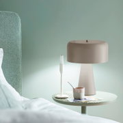Beige Tischlampe mit breitem, rundem Schirm auf rundem Nachttisch neben Kerzenhalter und Tasse, Innenraumaufnahme in seitlicher Perspektive