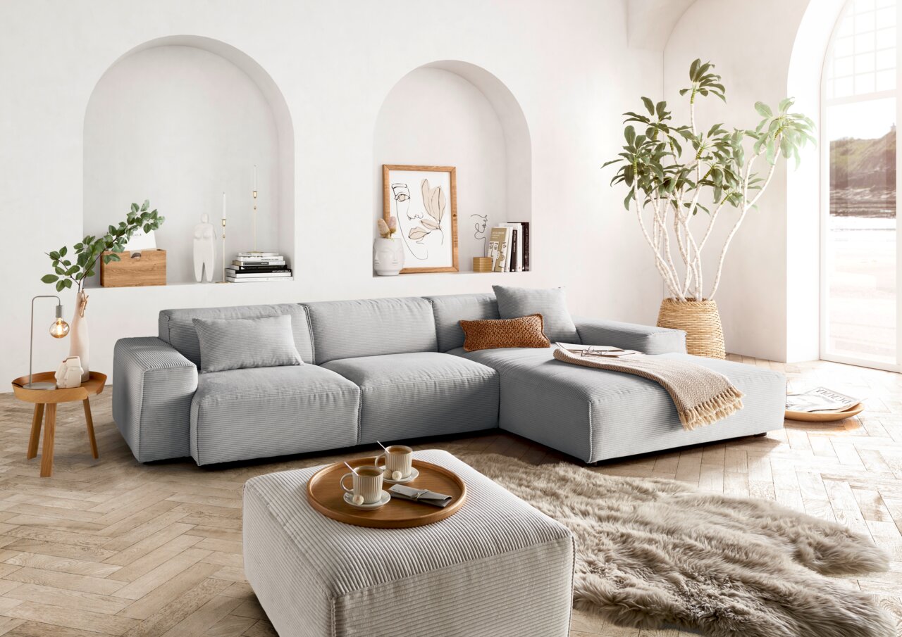 Lounge Handmade Ecksofa LH-EDINA Moderne graue Rundecke in einem hellen Wohnzimmer mit großen Fenstern, Holzboden und stilvoller Dekoration. Perspektive von vorne links.