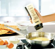 Hand hält Crepes- und Pfannkuchen-Mix-Flasche über einer Pfanne, aus der der Teig in die Pfanne fließt. Im Hintergrund sind Eier, Mehl und fertige Crepes zu sehen.