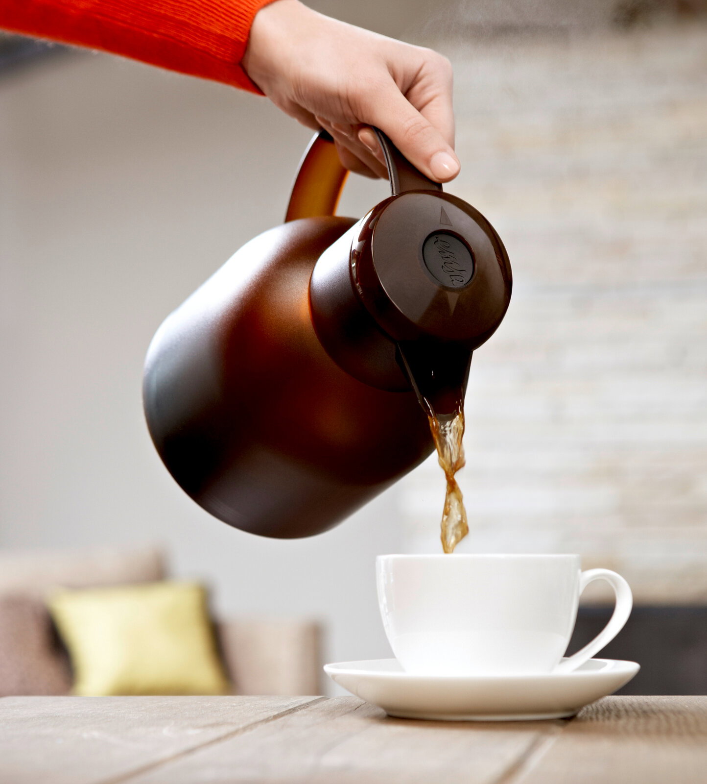 Eine Hand gießt Kaffee aus einer braunen Isolierkanne Samba QP 1,0l in eine weiße Tasse, seitliche Perspektive.