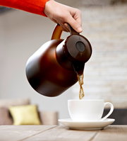 Eine Hand gießt Kaffee aus einer braunen Isolierkanne Samba QP 1,0l in eine weiße Tasse, seitliche Perspektive.