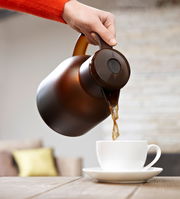 Eine Hand gießt Kaffee aus einer braunen Isolierkanne Samba QP 1,0l in eine weiße Tasse, seitliche Perspektive.