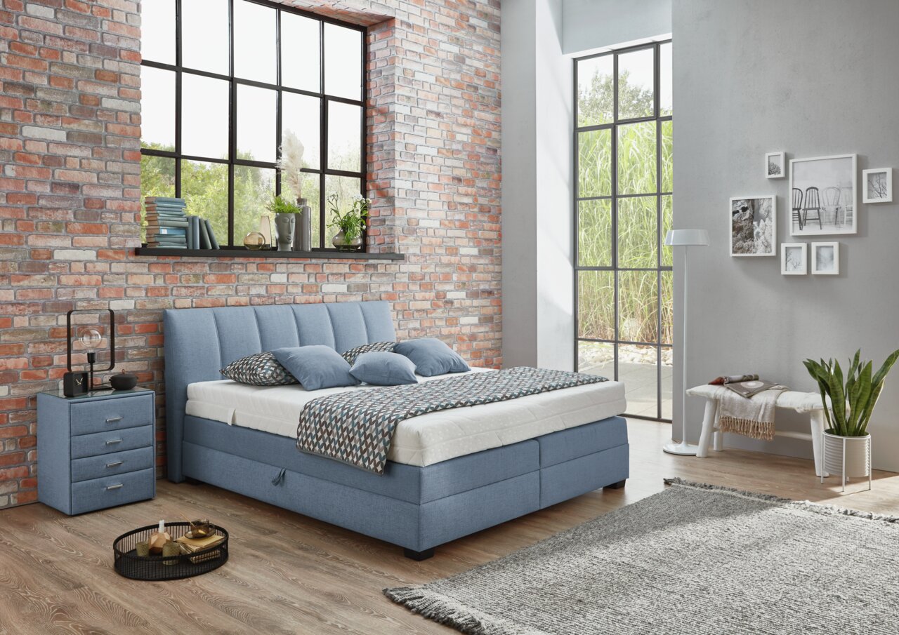 Boxspringbett in einem modernen Schlafzimmer mit Backsteinwand und großen Fenstern, seitliche Perspektive