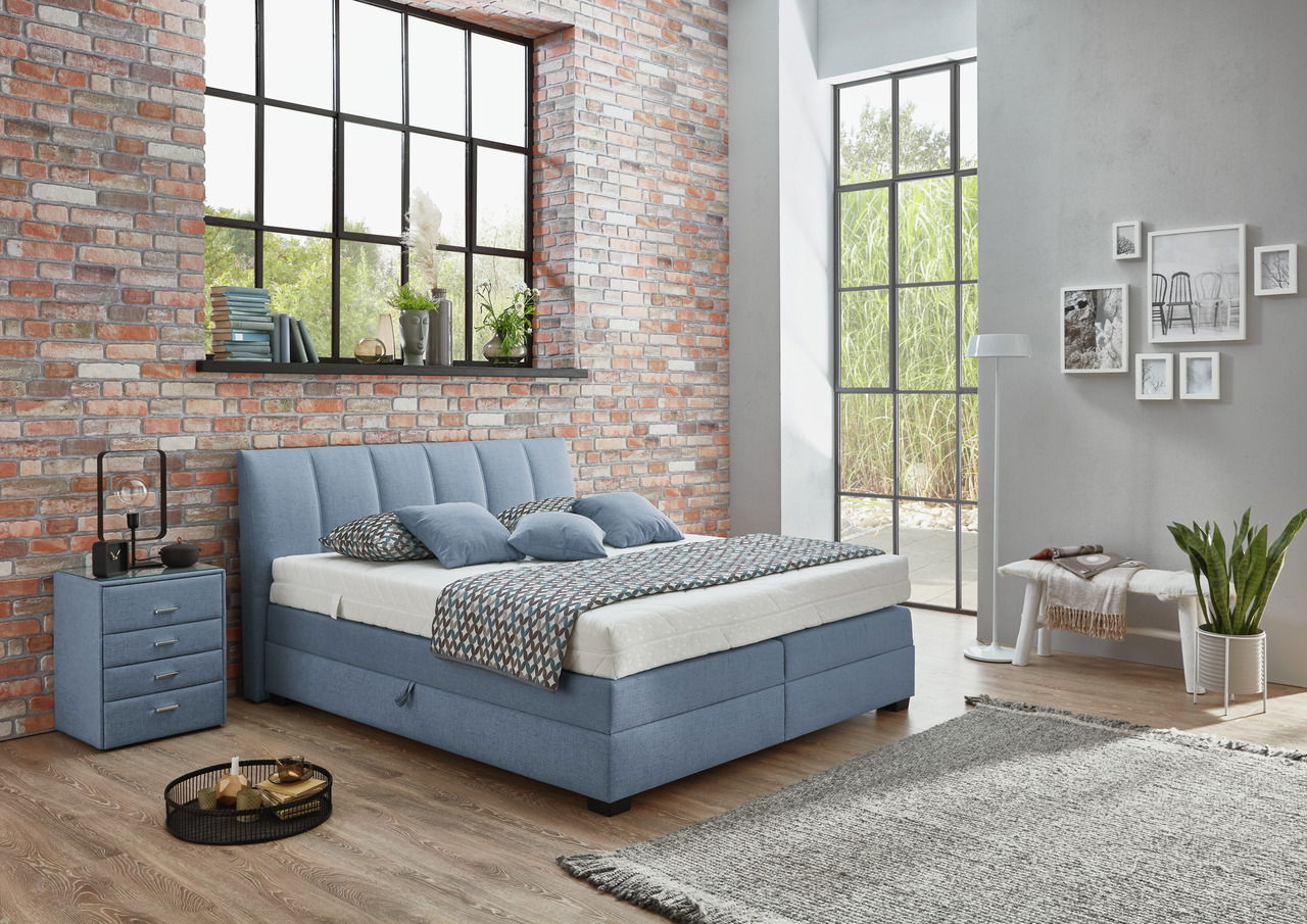 Boxspringbett in einem modernen Schlafzimmer mit Backsteinwand und großen Fenstern, seitliche Perspektive