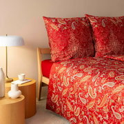 Rote Bettwäsche mit Paisley-Muster auf einem Bett, seitliche Perspektive mit Nachttisch und Lampe.