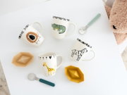 Vier liebevoll gestaltete Kinderbecher mit Löwen-, Krokodil-, Zebra- und Giraffenmotiv, zwei Löffel und zwei geometrische Schälchen auf einem weißen Tisch, aus der Vogelperspektive arrangiert.