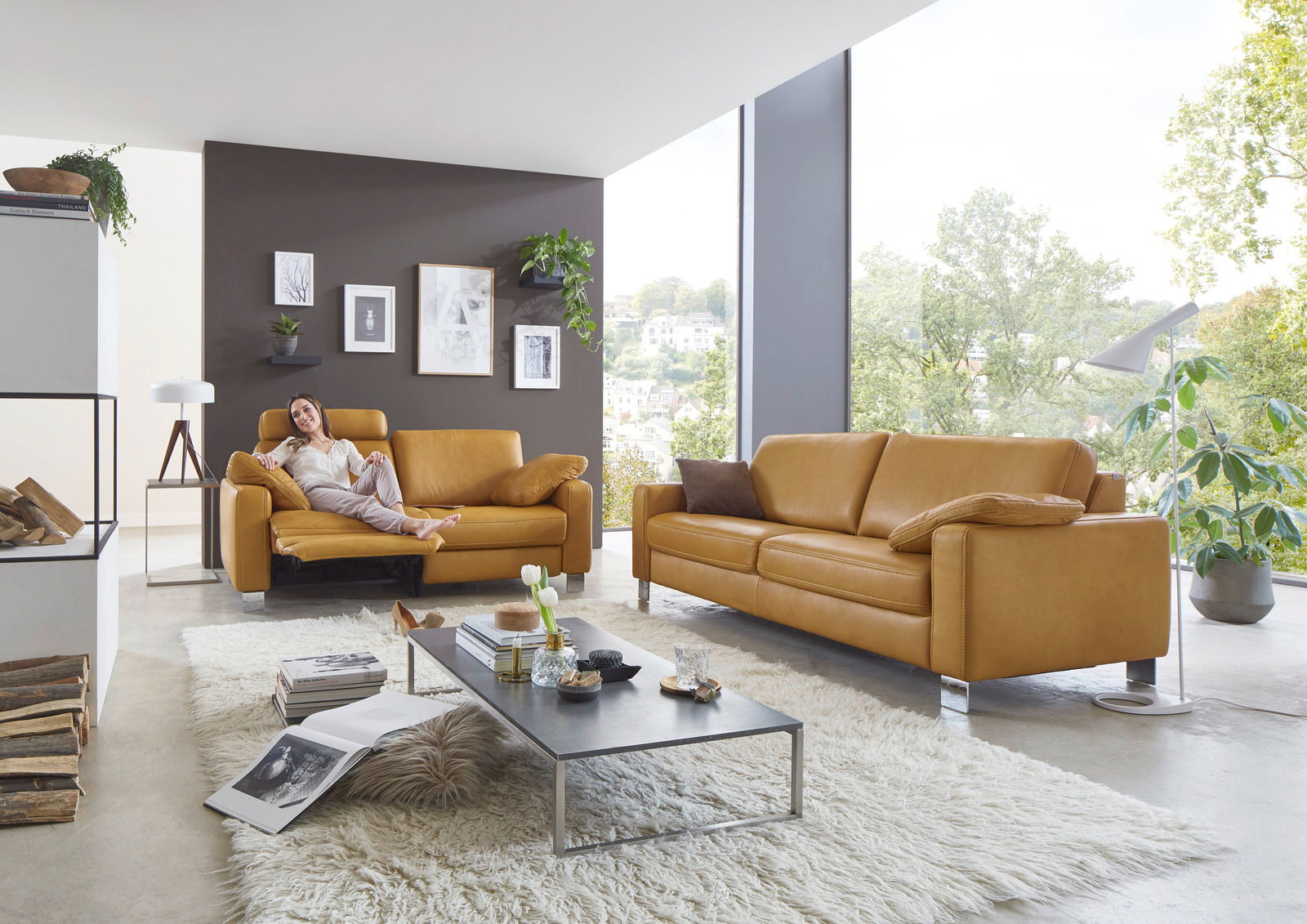 Modernes Wohnzimmer mit zwei senffarbenen Ledersofas, eines mit einer entspannten Person darauf, in einem hellen Raum mit großen Fenstern und grüner Aussicht.