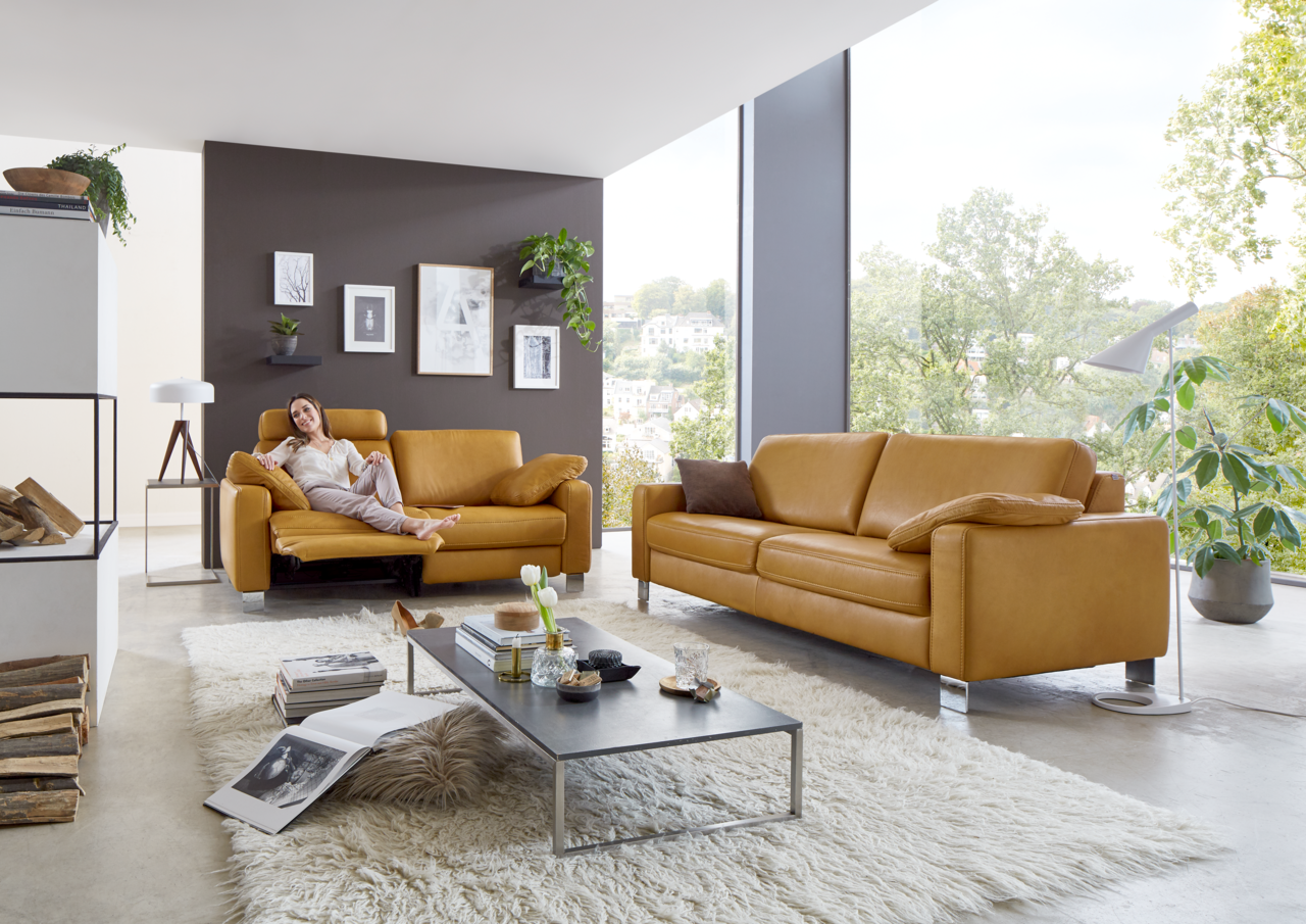 Modernes Wohnzimmer mit zwei senffarbenen Ledersofas, eines mit einer entspannten Person darauf, in einem hellen Raum mit großen Fenstern und grüner Aussicht.