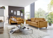Modernes Wohnzimmer mit zwei senffarbenen Ledersofas, eines mit einer entspannten Person darauf, in einem hellen Raum mit großen Fenstern und grüner Aussicht.