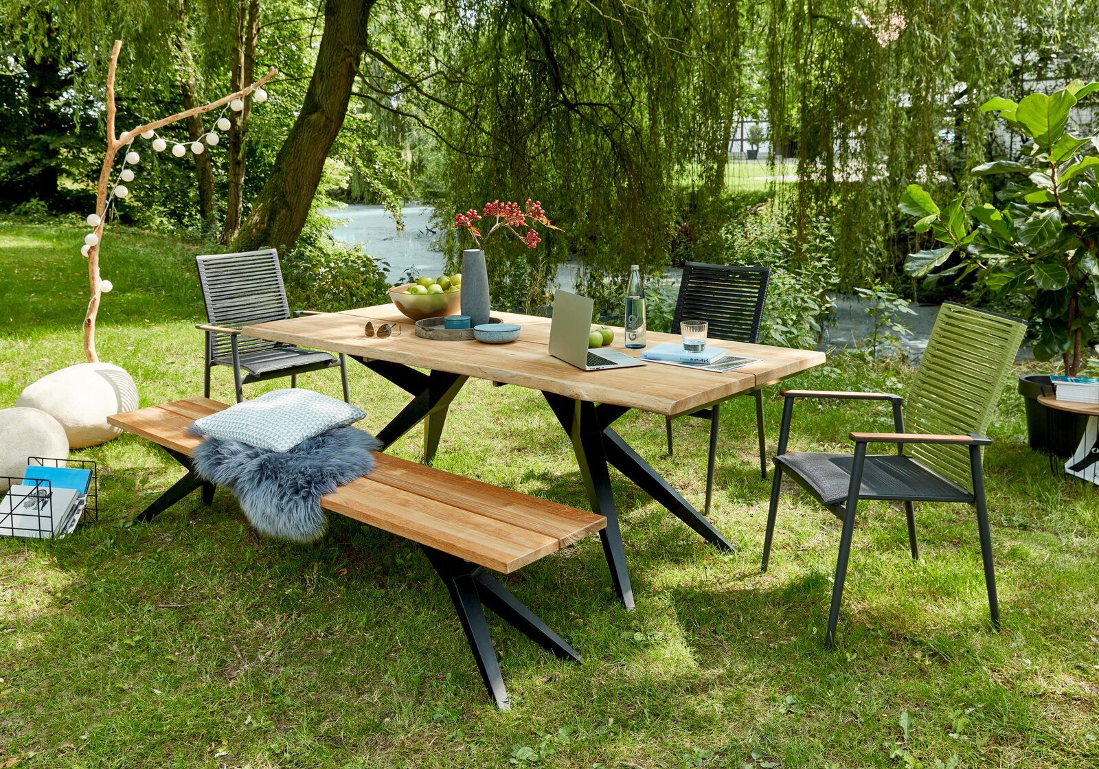 Gartenmöbel-Set bestehend aus einem Holztisch mit Metallbeinen, zwei Stühlen und einer Bank, aufgestellt auf einer grünen Wiese mit Blick auf einen Teich im Hintergrund.