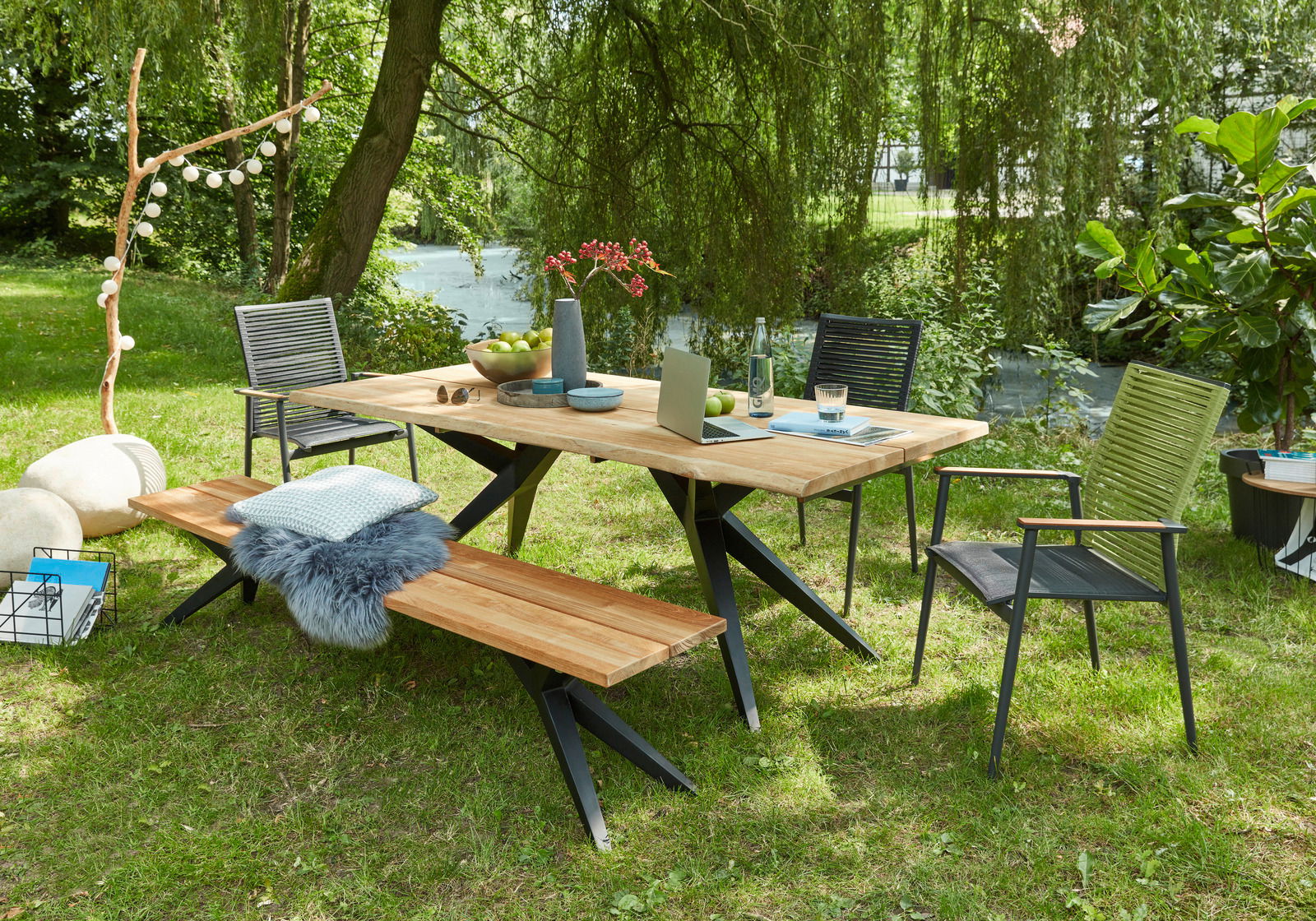 Gartenmöbel-Set bestehend aus einem Holztisch mit Metallbeinen, zwei Stühlen und einer Bank, aufgestellt auf einer grünen Wiese mit Blick auf einen Teich im Hintergrund.