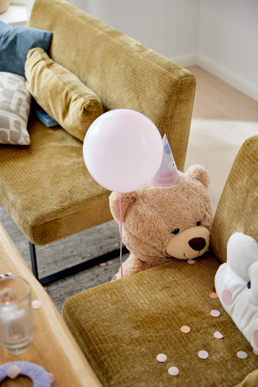 Gemütliche Wohnzimmerbank in senfgelber Farbe mit Kissen, einem Teddybär mit Partyhut und Luftballon, aus seitlicher Perspektive fotografiert.