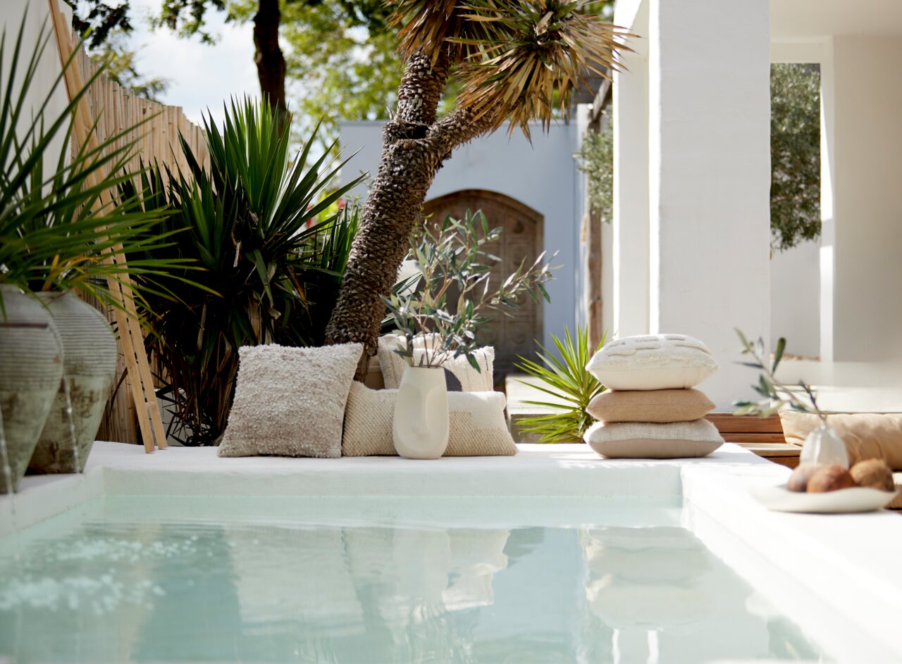 Ein stilvolles Kissen aus Bouclé-Stoff in einer entspannten Outdoor-Umgebung neben einem Pool, umgeben von grünen Pflanzen und einem modernen Dekor.