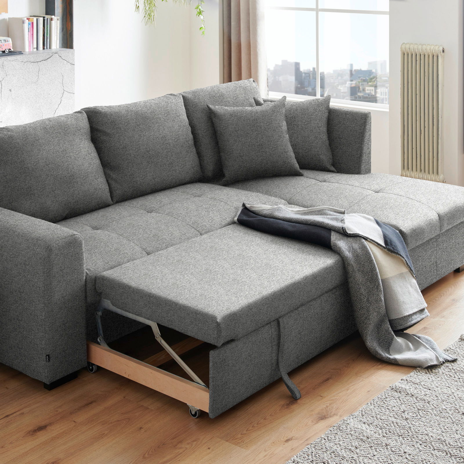 Graues Ecksofa mit ausziehbarem Bettkasten, dekoriert mit Kissen und einer Decke, in einem modernen Wohnzimmer mit Holzboden, seitliche Perspektive.