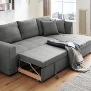 Graues Ecksofa mit ausziehbarem Bettkasten, dekoriert mit Kissen und einer Decke, in einem modernen Wohnzimmer mit Holzboden, seitliche Perspektive.