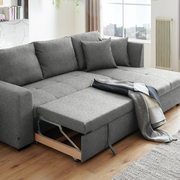 Graues Ecksofa mit ausziehbarem Bettkasten, dekoriert mit Kissen und einer Decke, in einem modernen Wohnzimmer mit Holzboden, seitliche Perspektive.