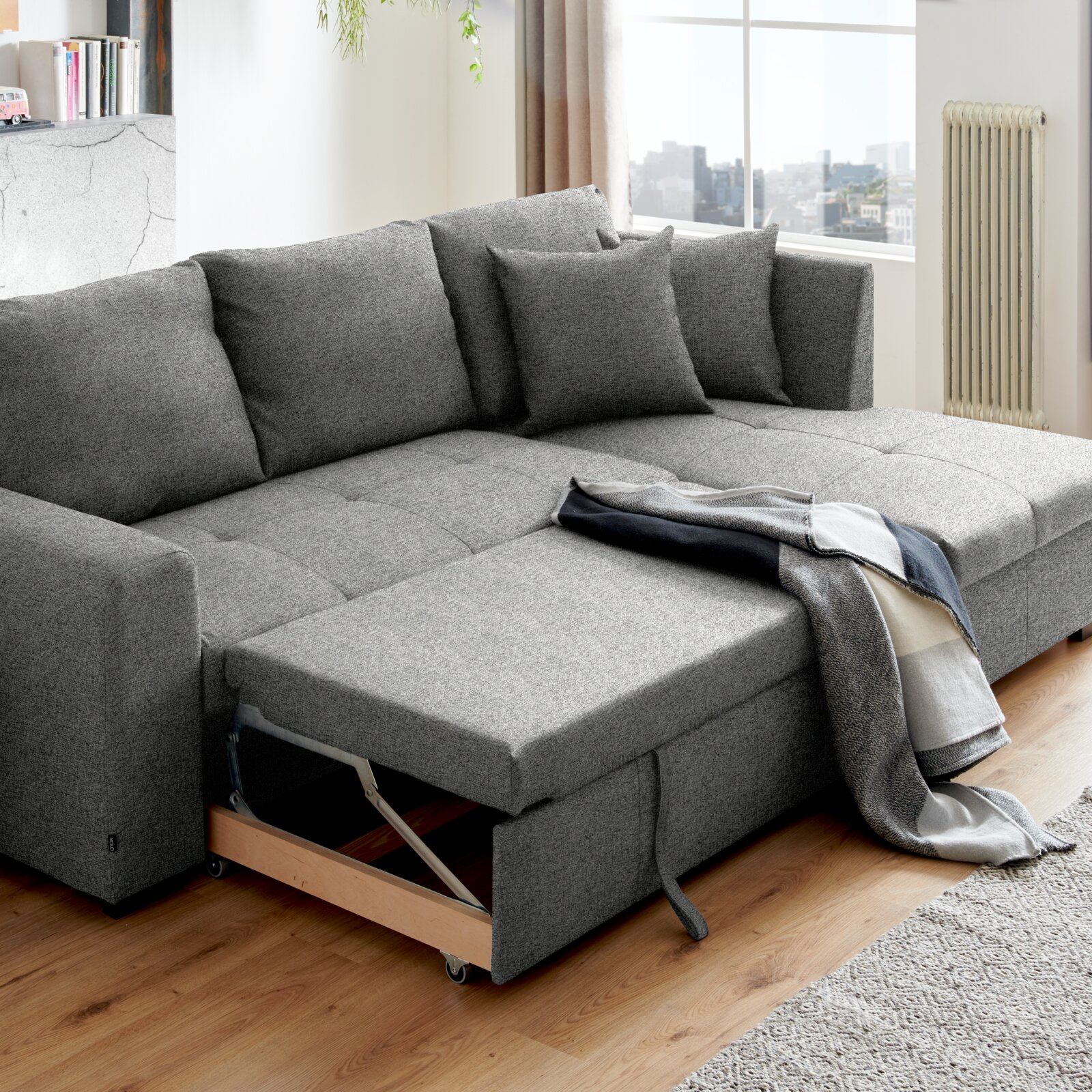 Graues Ecksofa mit ausziehbarem Bettkasten, dekoriert mit Kissen und einer Decke, in einem modernen Wohnzimmer mit Holzboden, seitliche Perspektive.