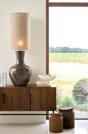 Leuchtende Tischlampe mit glänzendem, dunkelbraunem Keramikfuß und hohem, zylindrischem Leinenschirm, frontal auf einem Holz-Sideboard neben einem großen Fenster inszeniert