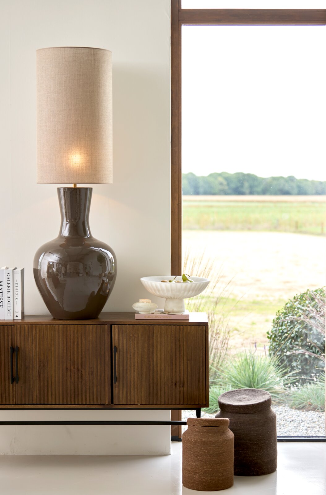 Leuchtende Tischlampe mit glänzendem, dunkelbraunem Keramikfuß und hohem, zylindrischem Leinenschirm, frontal auf einem Holz-Sideboard neben einem großen Fenster inszeniert