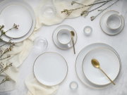 Draufsicht auf ein elegant gedecktes Tisch-Arrangement mit weißen, geriffelten Porzellantellern und -tassen mit Goldrand, goldfarbenen Löffeln, Glasuntersetzern, kleinen Kerzen im Glas, hellem Stoff und zarten Blütenzweigen auf heller Oberfläche