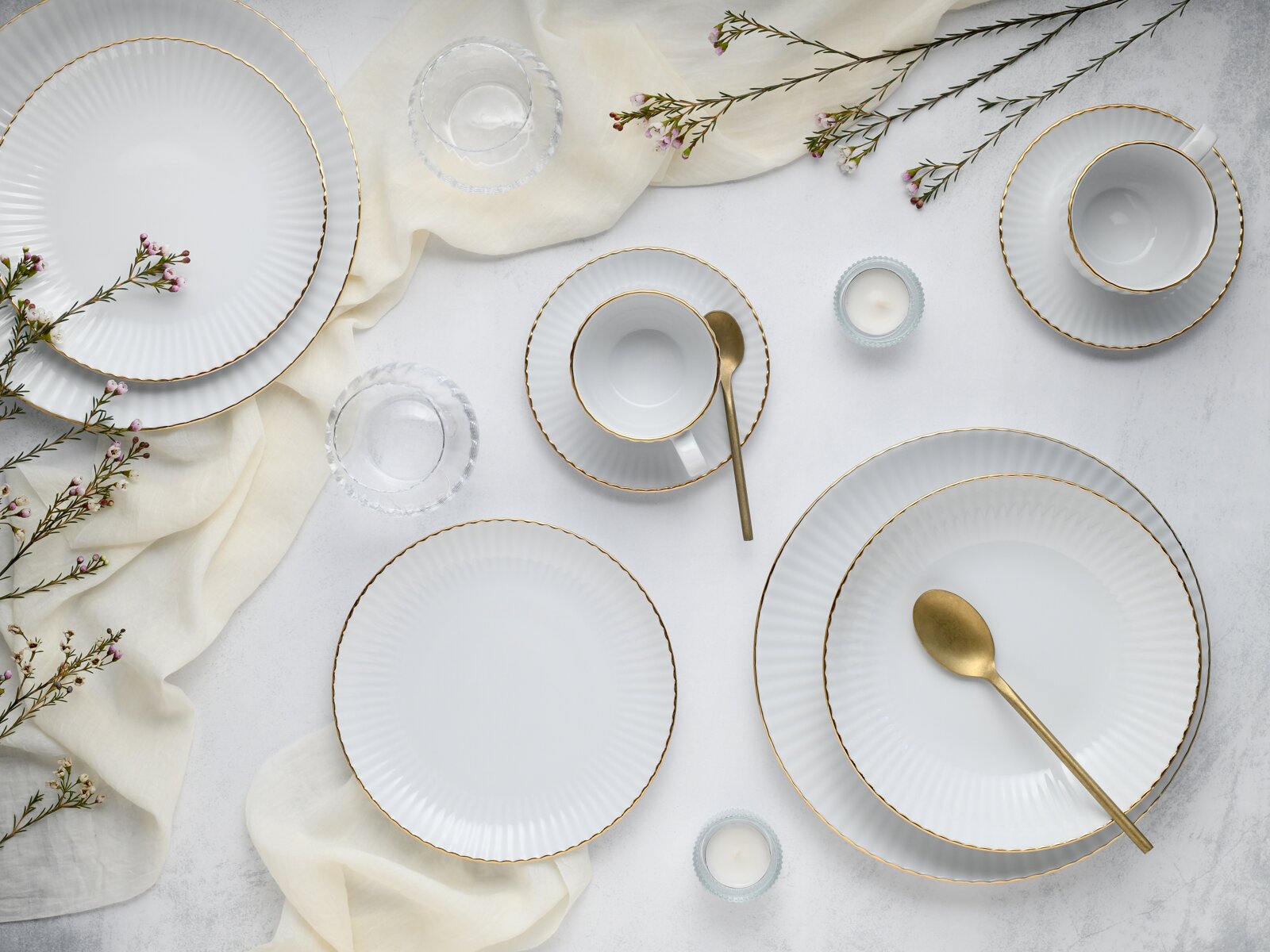 Draufsicht auf ein elegant gedecktes Tisch-Arrangement mit weißen, geriffelten Porzellantellern und -tassen mit Goldrand, goldfarbenen Löffeln, Glasuntersetzern, kleinen Kerzen im Glas, hellem Stoff und zarten Blütenzweigen auf heller Oberfläche