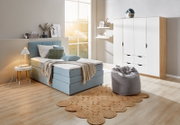 Gemütliches Schlafzimmer mit einem blauen Boxspringbett, das von der Seite betrachtet wird. Das Bett ist mit Kissen und einer Decke dekoriert, daneben steht ein kleiner runder Tisch mit einer Lampe. Im Hintergrund sind ein Schrank und ein Fenster zu sehen