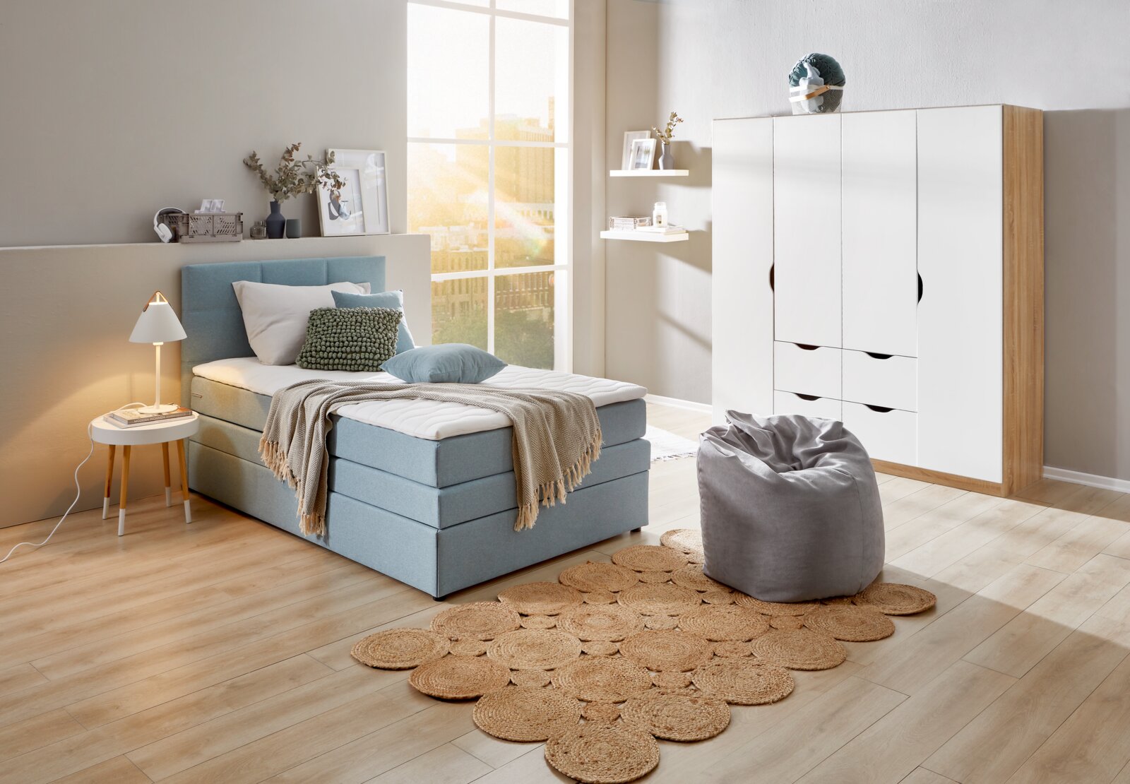 Gemütliches Schlafzimmer mit einem blauen Boxspringbett, das von der Seite betrachtet wird. Das Bett ist mit Kissen und einer Decke dekoriert, daneben steht ein kleiner runder Tisch mit einer Lampe. Im Hintergrund sind ein Schrank und ein Fenster zu sehen