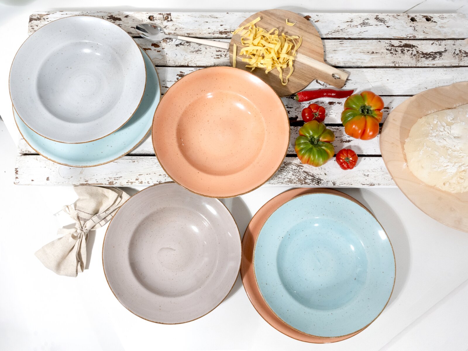 Pastellfarbene Keramik-Speiseteller in Blau, Rosa und Grau mit gesprenkelter Glasur auf rustikalem Holztisch, Draufsicht (Flatlay) mit Nudeln, Tomaten und Teig