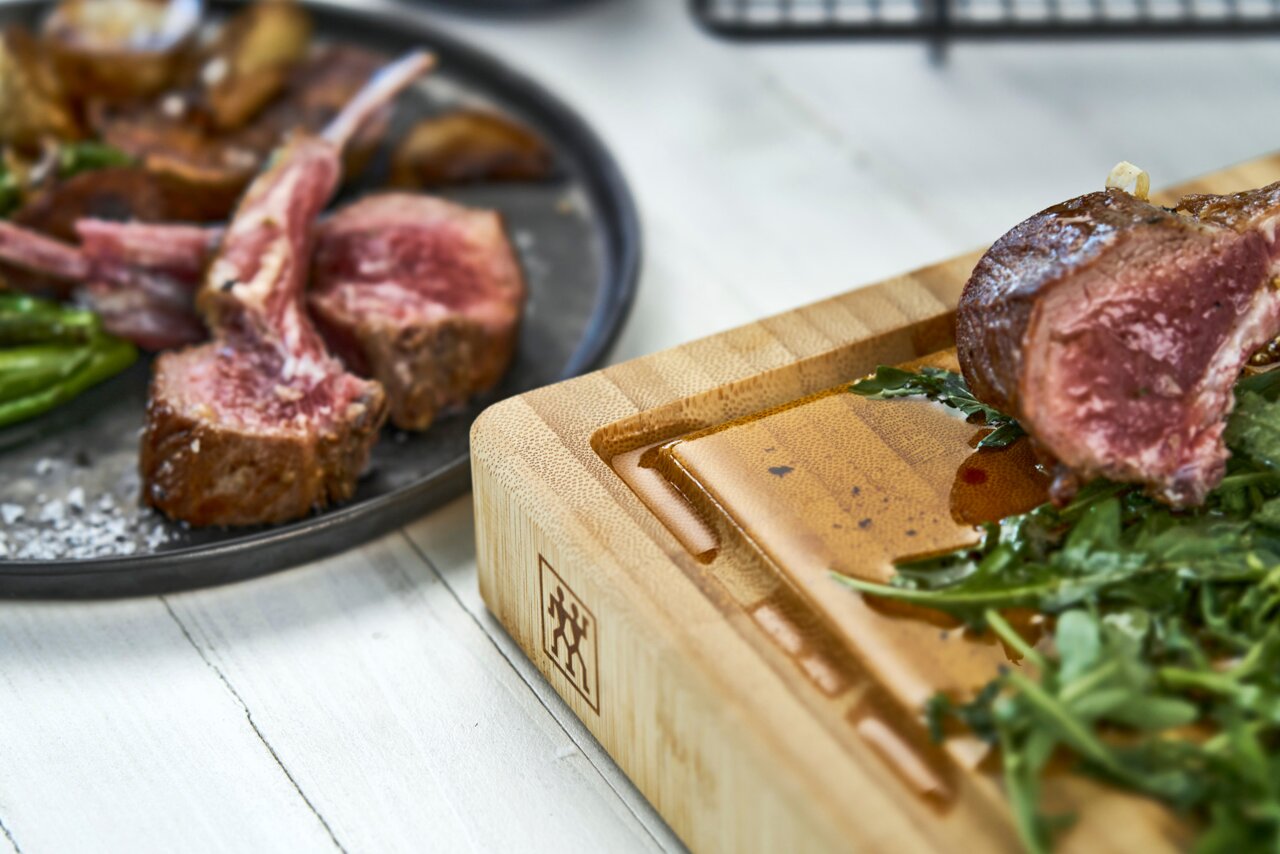 Nahaufnahme eines Bambus-Schneidbretts der Marke ZWILLING mit saftigem Fleisch und Kräutern, seitliche Perspektive.