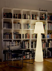 Leuchtende, moderne Stehleuchte in Weiß vor einem großen weißen Bücherregal, daneben ein schwarzer Freischwinger-Stuhl; Aufnahme aus frontaler Perspektive auf Augenhöhe im Wohnraum.