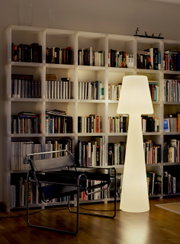 Leuchtende, moderne Stehleuchte in Weiß vor einem großen weißen Bücherregal, daneben ein schwarzer Freischwinger-Stuhl; Aufnahme aus frontaler Perspektive auf Augenhöhe im Wohnraum.
