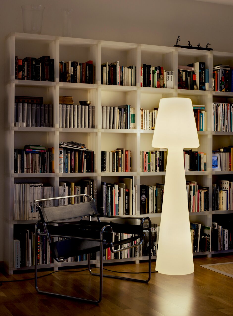 Een stralende, moderne staande lamp in wit voor een grote witte boekenkast, met daarnaast een zwarte sledestoel; foto vanuit een frontaal perspectief op ooghoogte in de woonkamer.