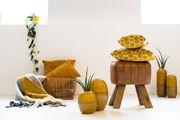Moderner Hocker aus braunem Leder mit Holzbeinen, im Vordergrund mit gelben Kissen und Vasen dekoriert, seitliche Perspektive.