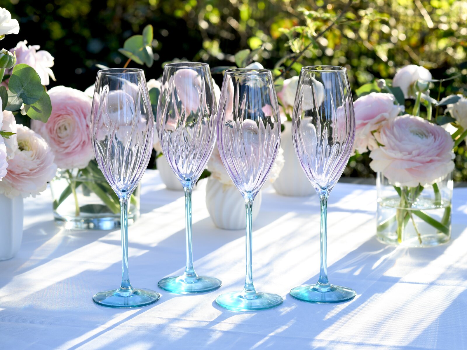 Vier elegante Sektgläser aus klarem, facettiertem Glas mit leicht blaugrün getöntem Stiel und Fuß auf weißer Tischdecke, im Hintergrund rosa Blumenarrangement im Garten; frontale Perspektive auf Augenhöhe.