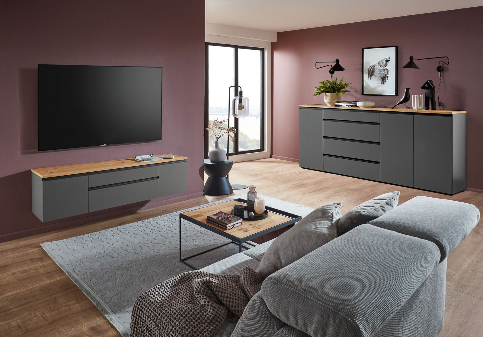 Modernes Wohnzimmer mit grauem TV-Lowboard und Sideboard, beide mit Holzoberfläche, vor einer dunkelroten Wand. Perspektive von der Seite.
