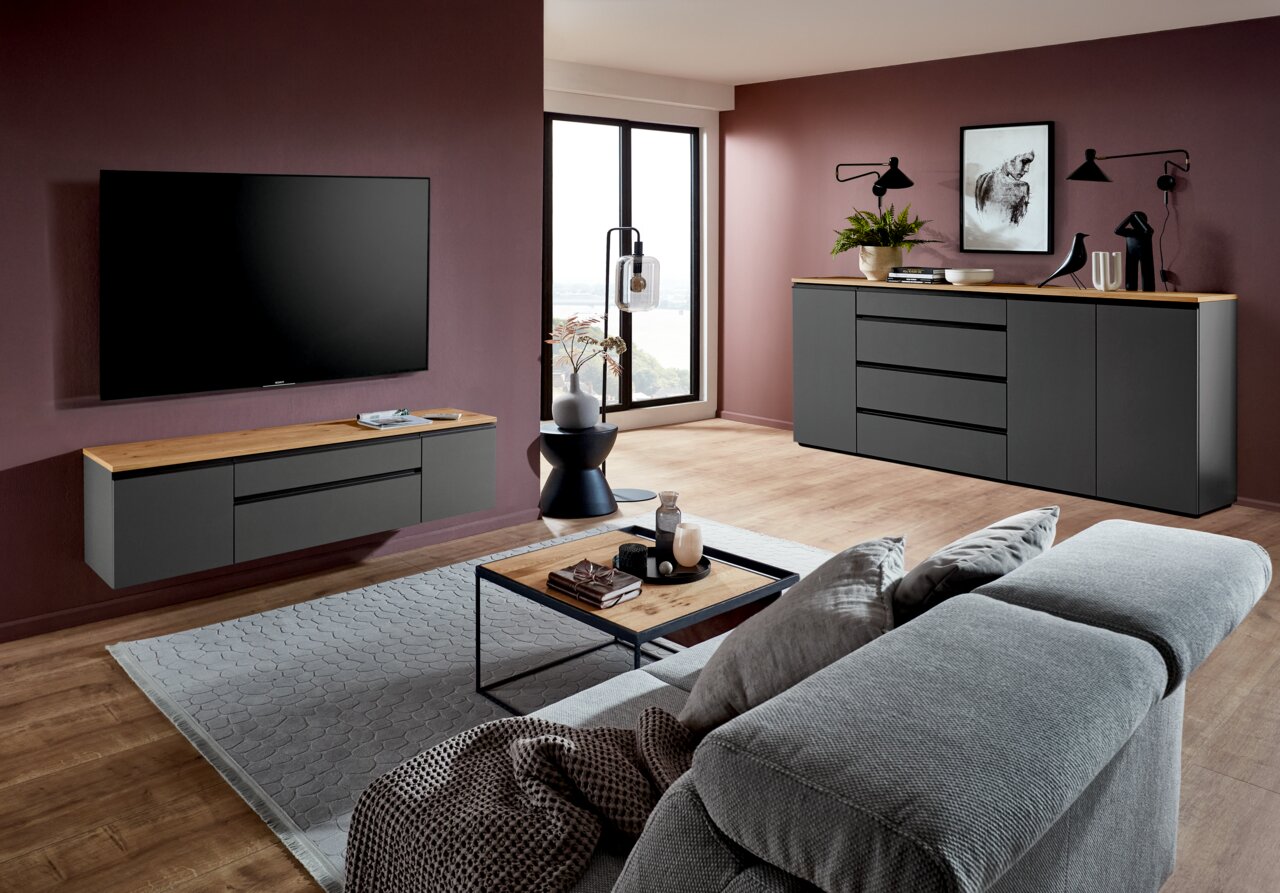 Modernes Wohnzimmer mit grauem TV-Lowboard und Sideboard, beide mit Holzoberfläche, vor einer dunkelroten Wand. Perspektive von der Seite.
