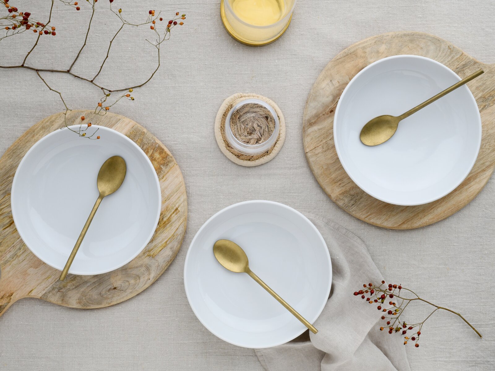 Draufsicht auf ein minimalistisches Tisch-Arrangement mit drei weißen Keramikschalen auf runden Holzbrettchen, jeweils mit goldfarbenem Löffel, auf hellem Leinentuch mit Beerenzweigen und einem Glas mit gelber Flüssigkeit.